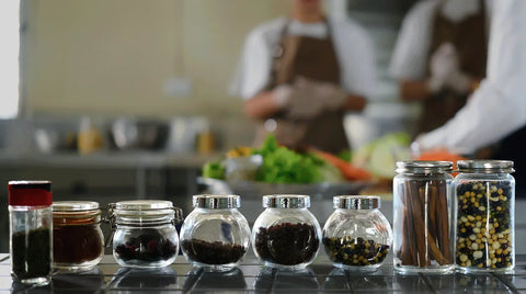 A row of clear glass jars with silver lids containing various whole spices and herbs, arranged on a reflective dark countertop in a blurred kitchen setting with chefs in the background.