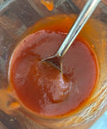 A close-up shot of a thick, glossy reddish-brown sauce or puree inside a clear container, with a metal spoon resting in it.