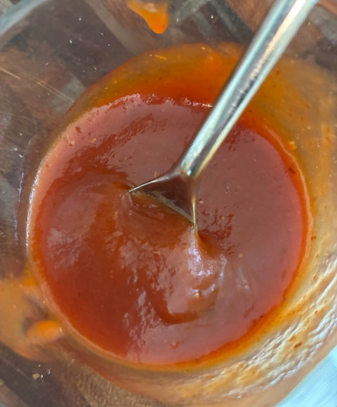 A close-up shot of a thick, glossy reddish-brown sauce or puree inside a clear container, with a metal spoon resting in it.